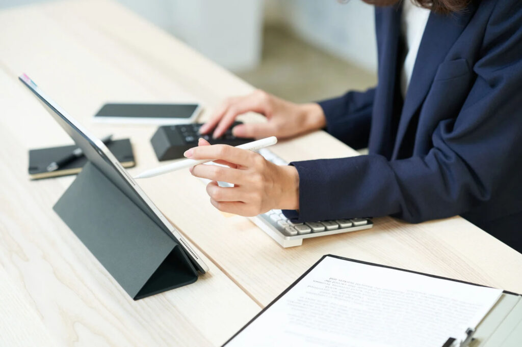 A person in a dark jacket using a tablet with a stylus while sitting at a desk. A calculator, notebook, and smartphone are visible on the desk.