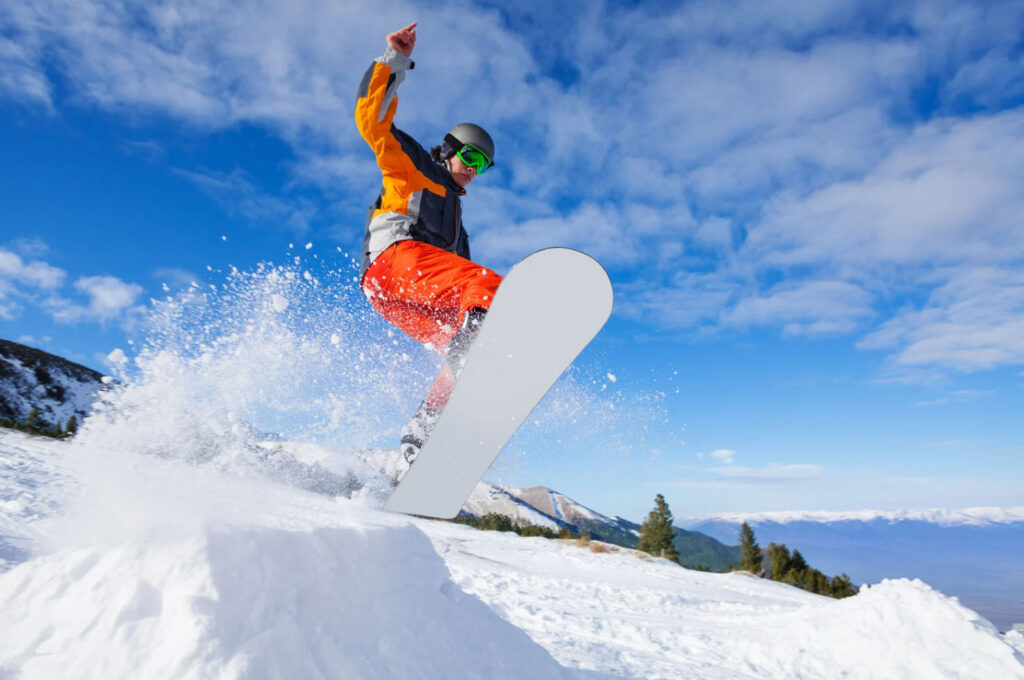 A snowboarder in orange pants and a grey jacket performing a trick on snowy slopes, with blue skies and mountains in the background.