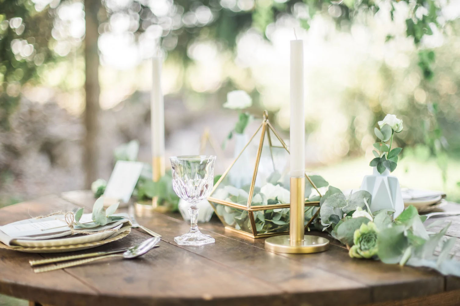 A beautifully arranged table setting with gold cutlery, a crystal glass, white candles in golden holders, and geometric vases with greenery and flowers, surrounded by a soft, natural background of blurred greenery.