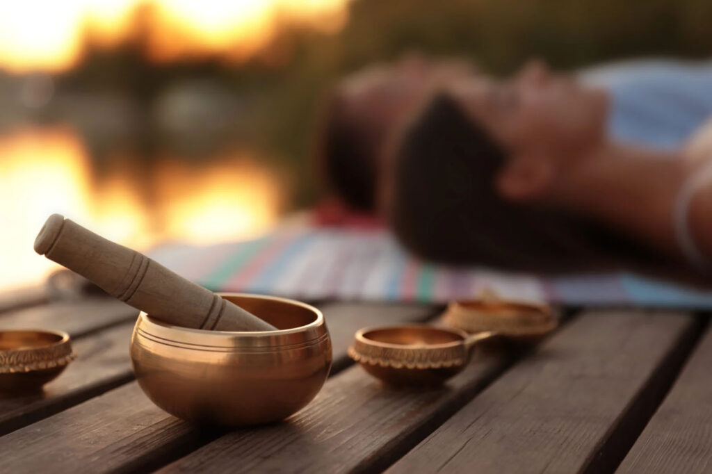 A close-up of a Tibetan singing bowl with a wooden mallet resting on it, placed on a wooden surface. In the background, a person is lying on a colorful mat by a lake, blurred out with warm sunlight in the distance.