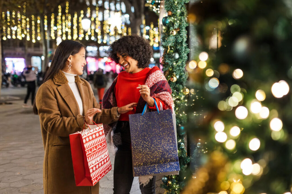 Two women exchanging gifts in a festive outdoor setting with holiday lights and decorations in the background, both smiling and holding colorful gift bags.