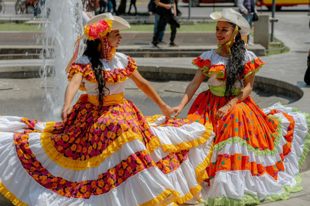 Two women in colorful floral dresses dancing joyfully by a fountain, smiling and wearing wide-brimmed hats, with people walking in the background.