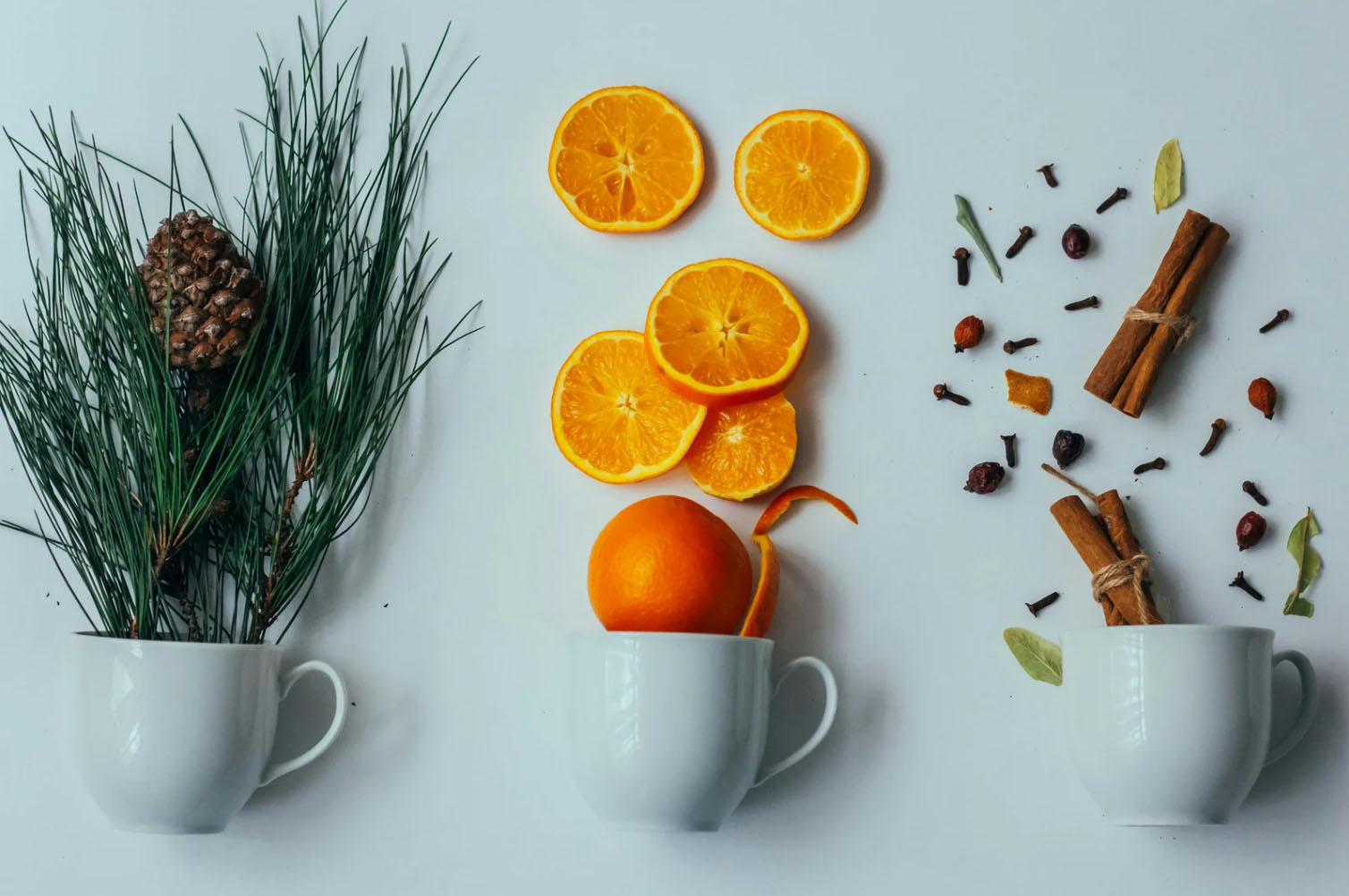 A creative arrangement featuring three white cups, each filled with different natural items: the first cup contains pine branches and a pine cone, the second holds an orange and orange slices, and the third has cinnamon sticks, cloves, and dried leaves scattered around.