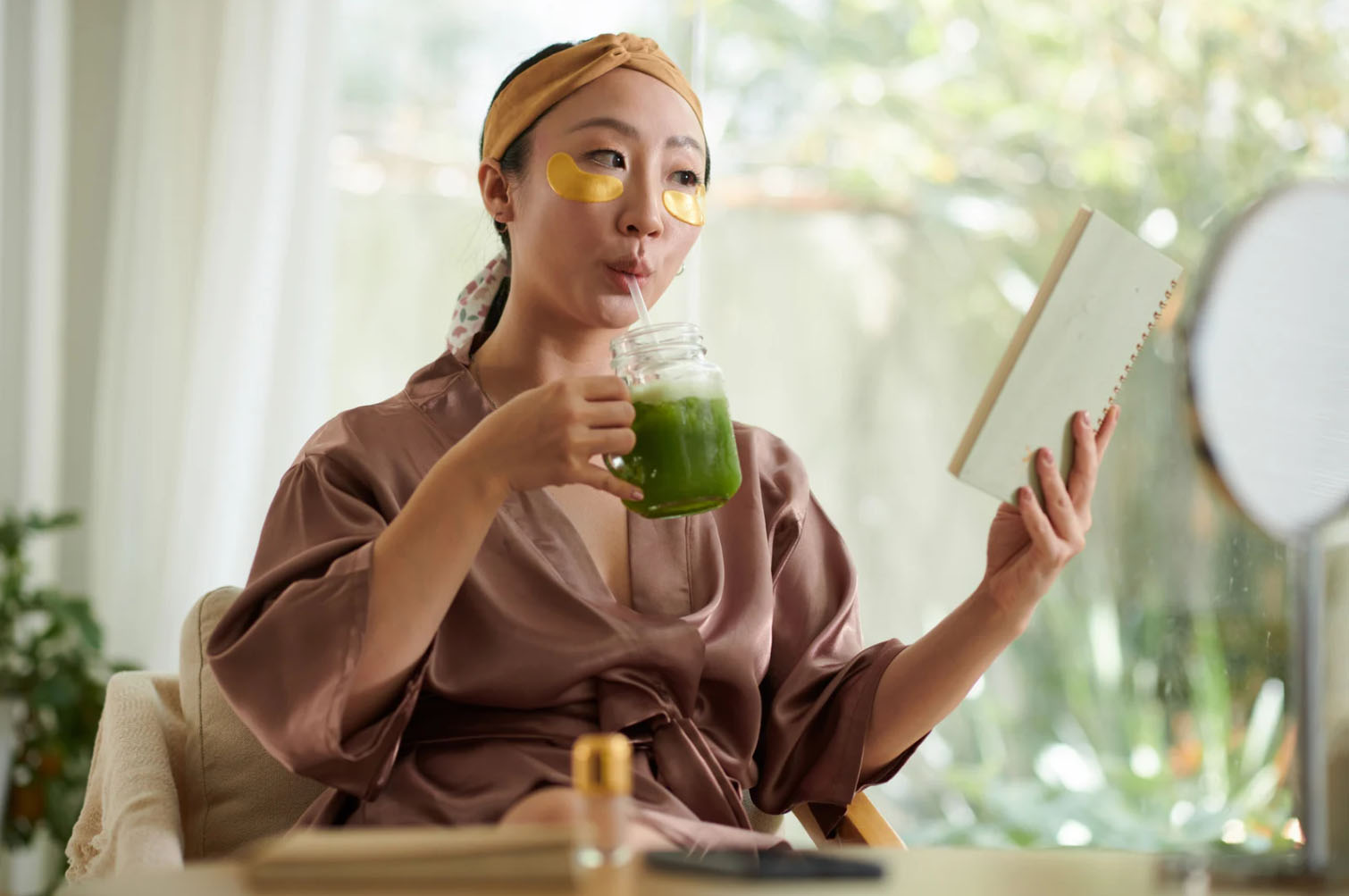 A woman in a satin robe enjoys a healthy green drink while reading a notebook, with under-eye patches on her face for relaxation, in a serene indoor setting.