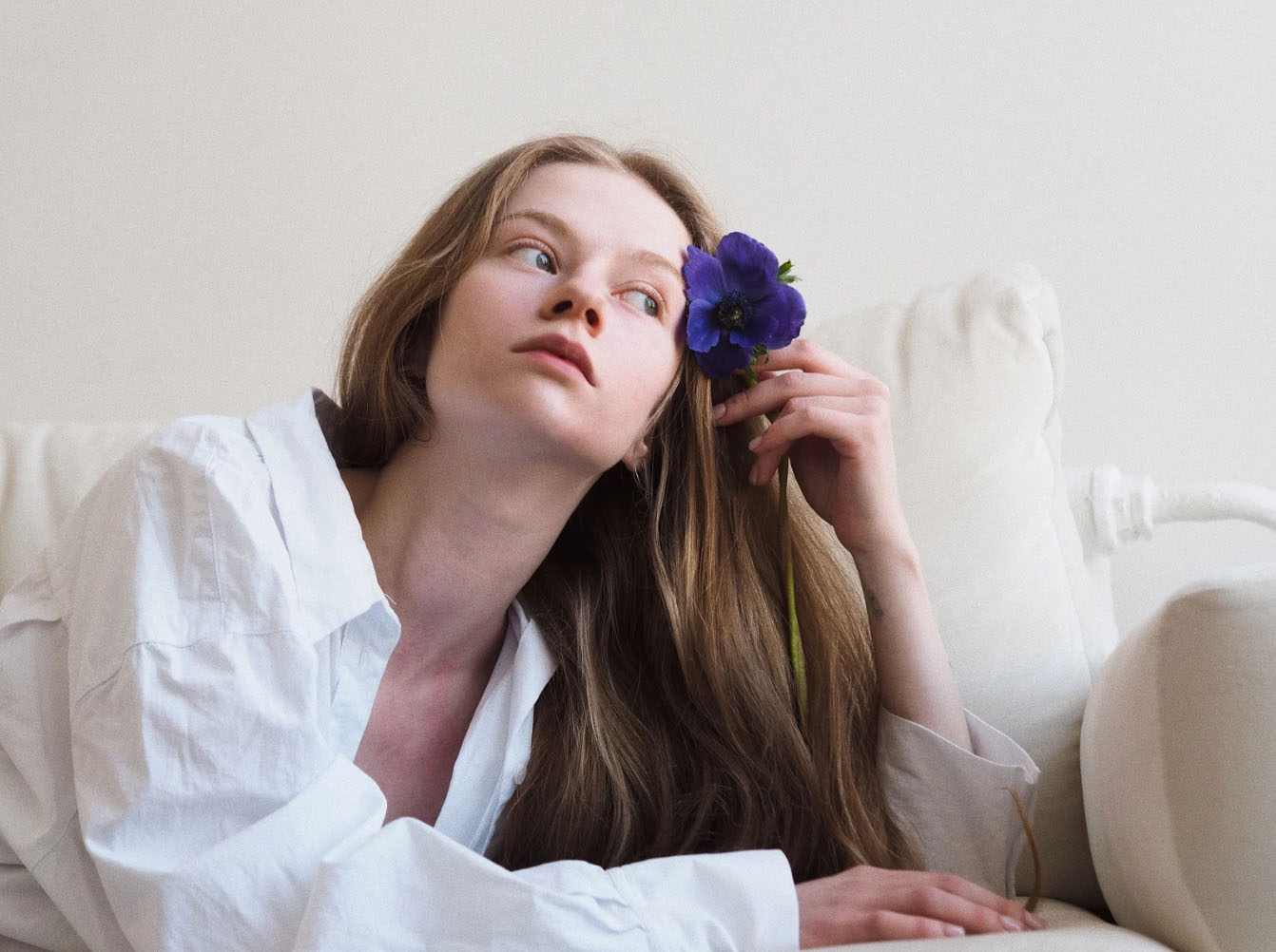 A woman in a white shirt lies on a white couch, gently holding a purple flower near her face, with a thoughtful expression as she gazes to the side.