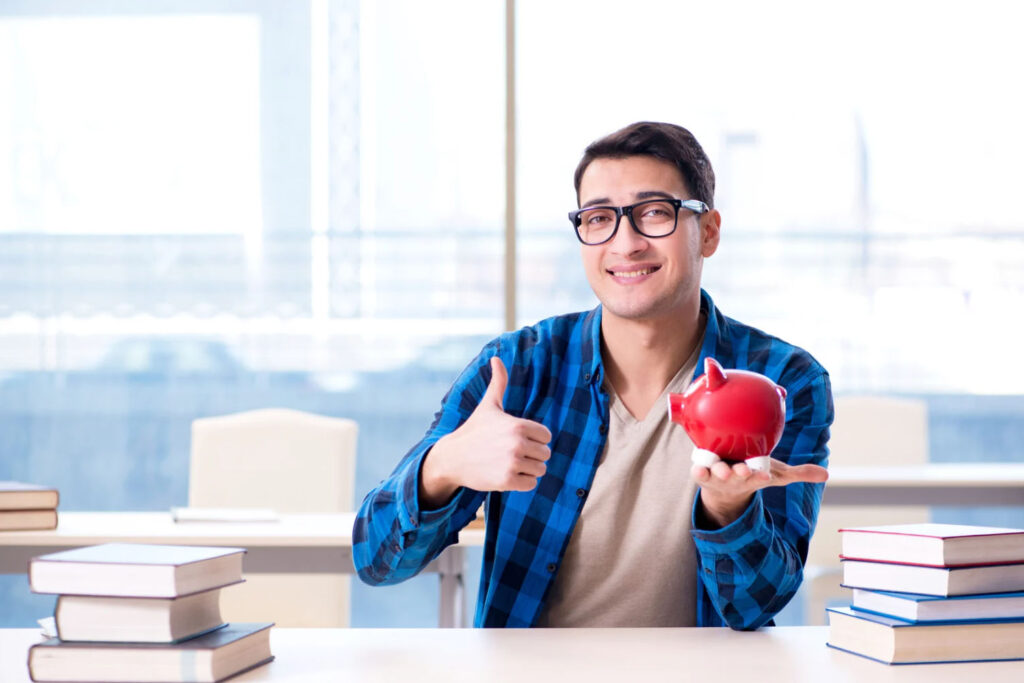 A young man smiles and gives a thumbs-up while holding a red piggy bank, with books on the table in a bright, modern office setting.