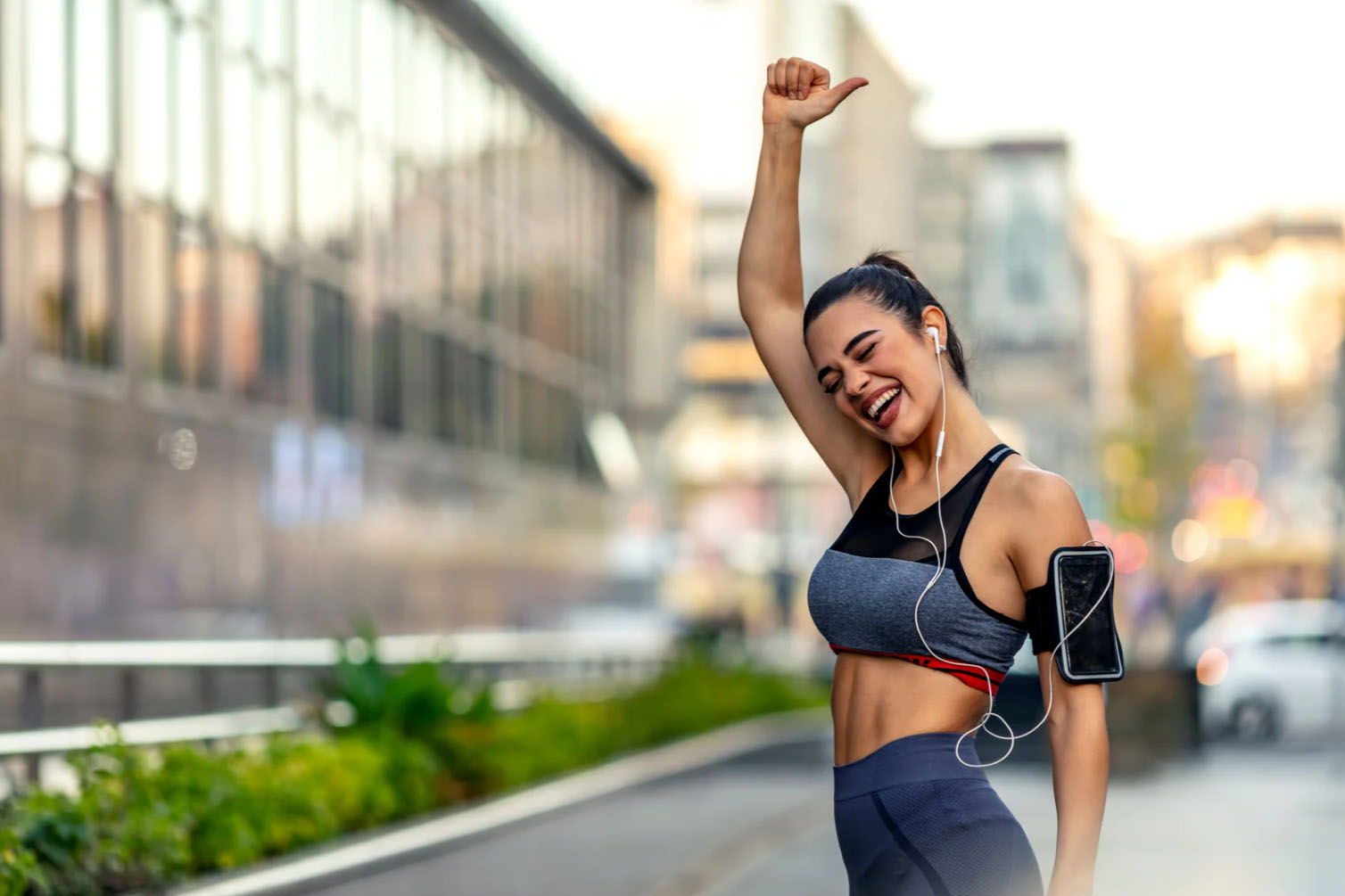 A young woman in athletic wear, smiling and celebrating with her arm raised in victory while listening to music outdoors in an urban setting.