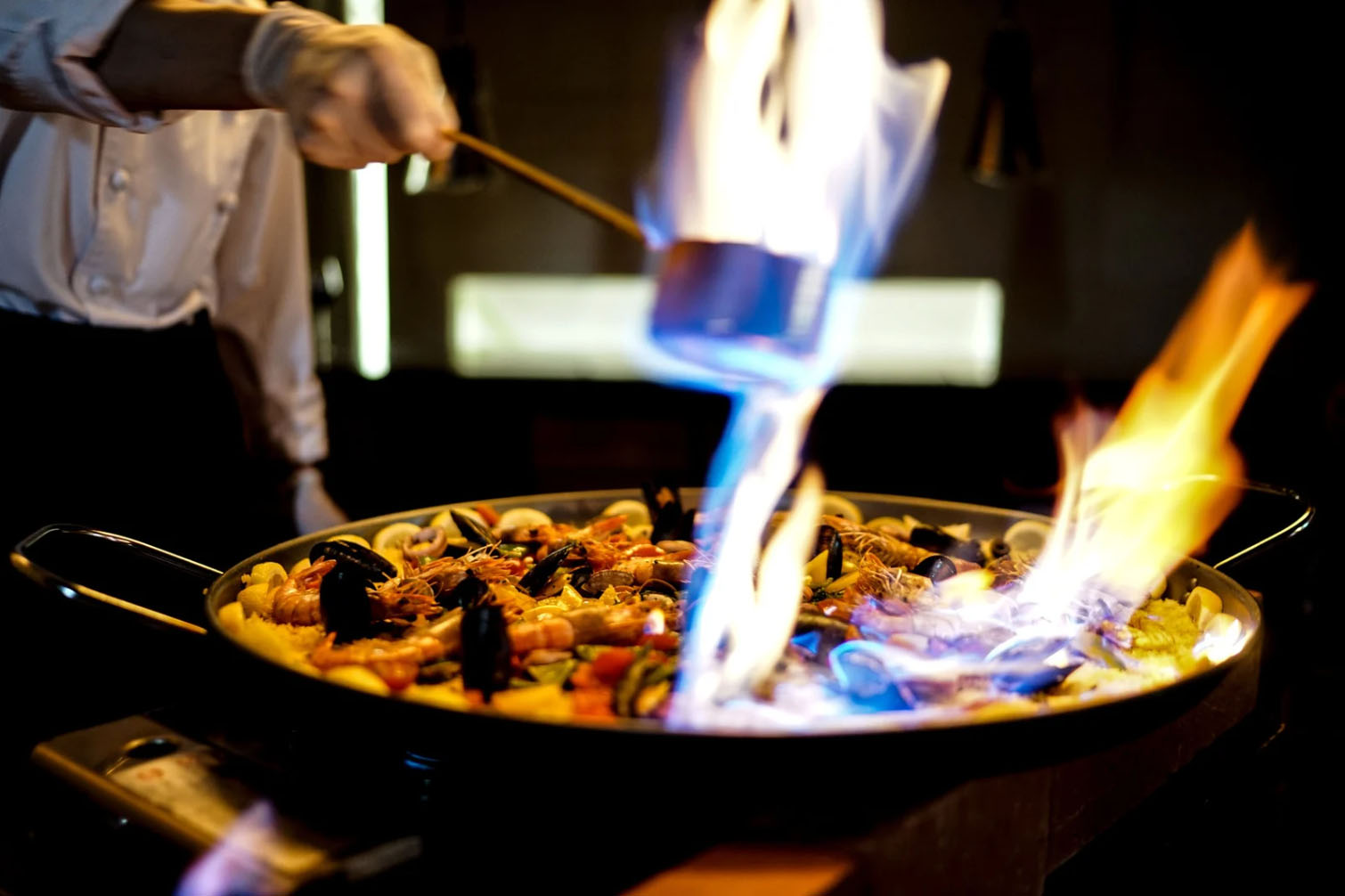 A chef flames a seafood paella dish with a torch, adding dramatic flair as the fire rises above the ingredients in the pan.