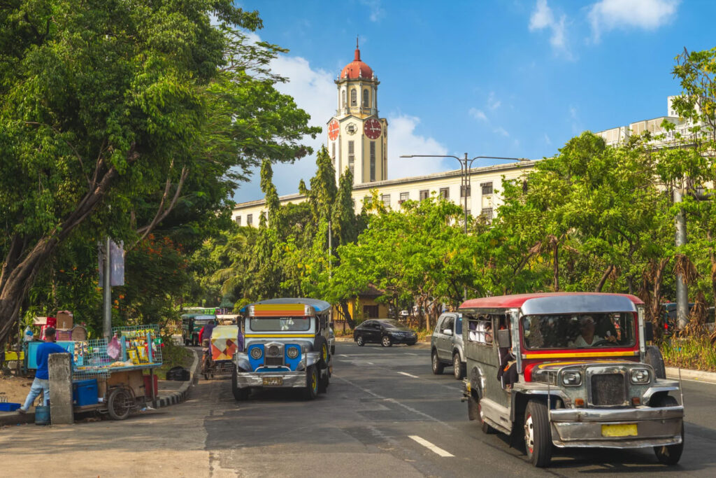 A busy street scene in Manila with colorful jeepneys, vendors selling goods, and the iconic clock tower of Manila City Hall in the background, framed by trees and blue skies.