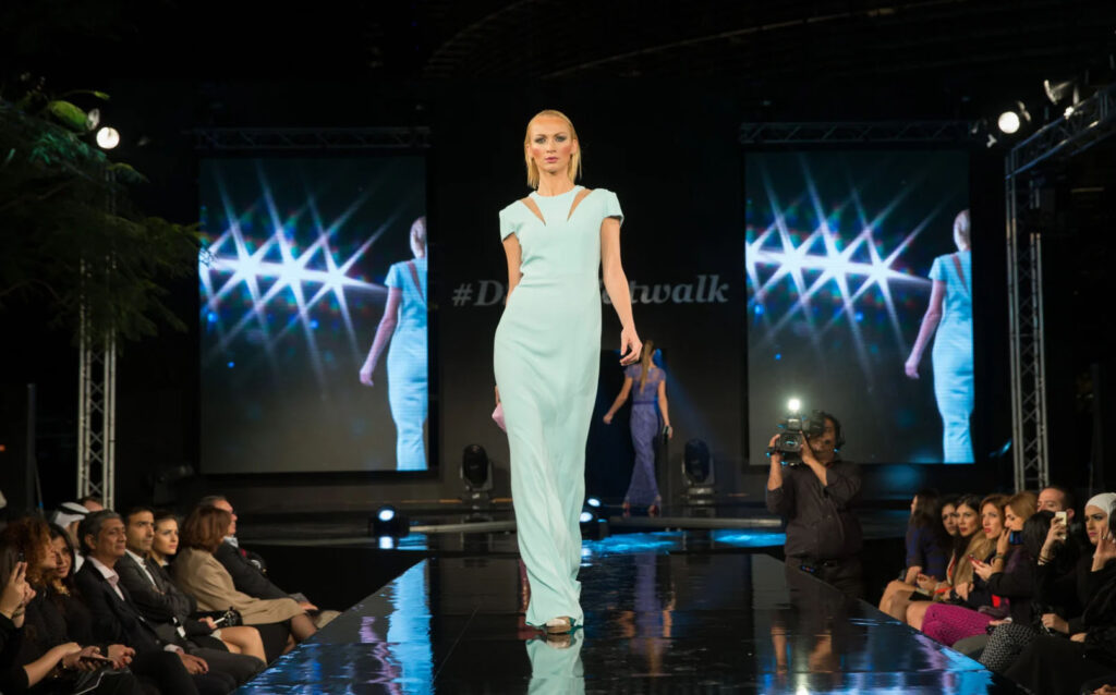 A model walks down the runway at a fashion show, wearing a light blue dress, while photographers capture the moment and an audience watches from the front row.