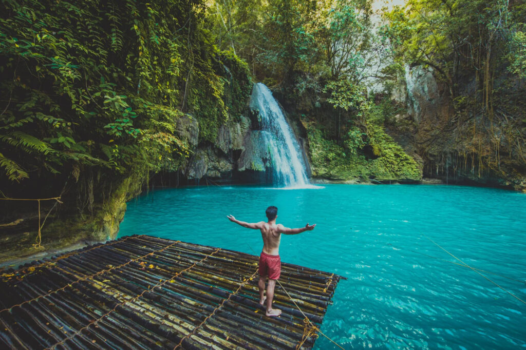 A person standing on a bamboo raft in front of a turquoise waterfall surrounded by lush greenery, with arms outstretched, enjoying the serene nature of the scene.