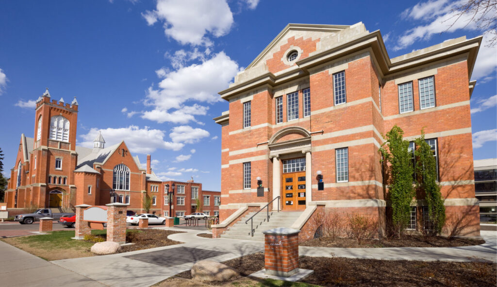 A beautiful historical building with brick architecture, featuring large windows, a set of stairs, and greenery in front, set against a clear blue sky.