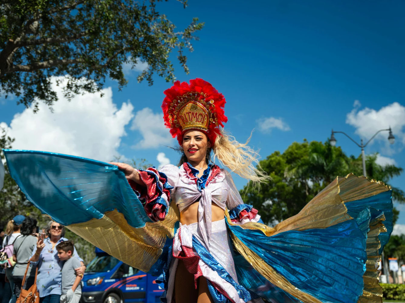 A woman dressed in a colorful carnival costume with a feathered headdress, dancing at a festival parade with people in the background.