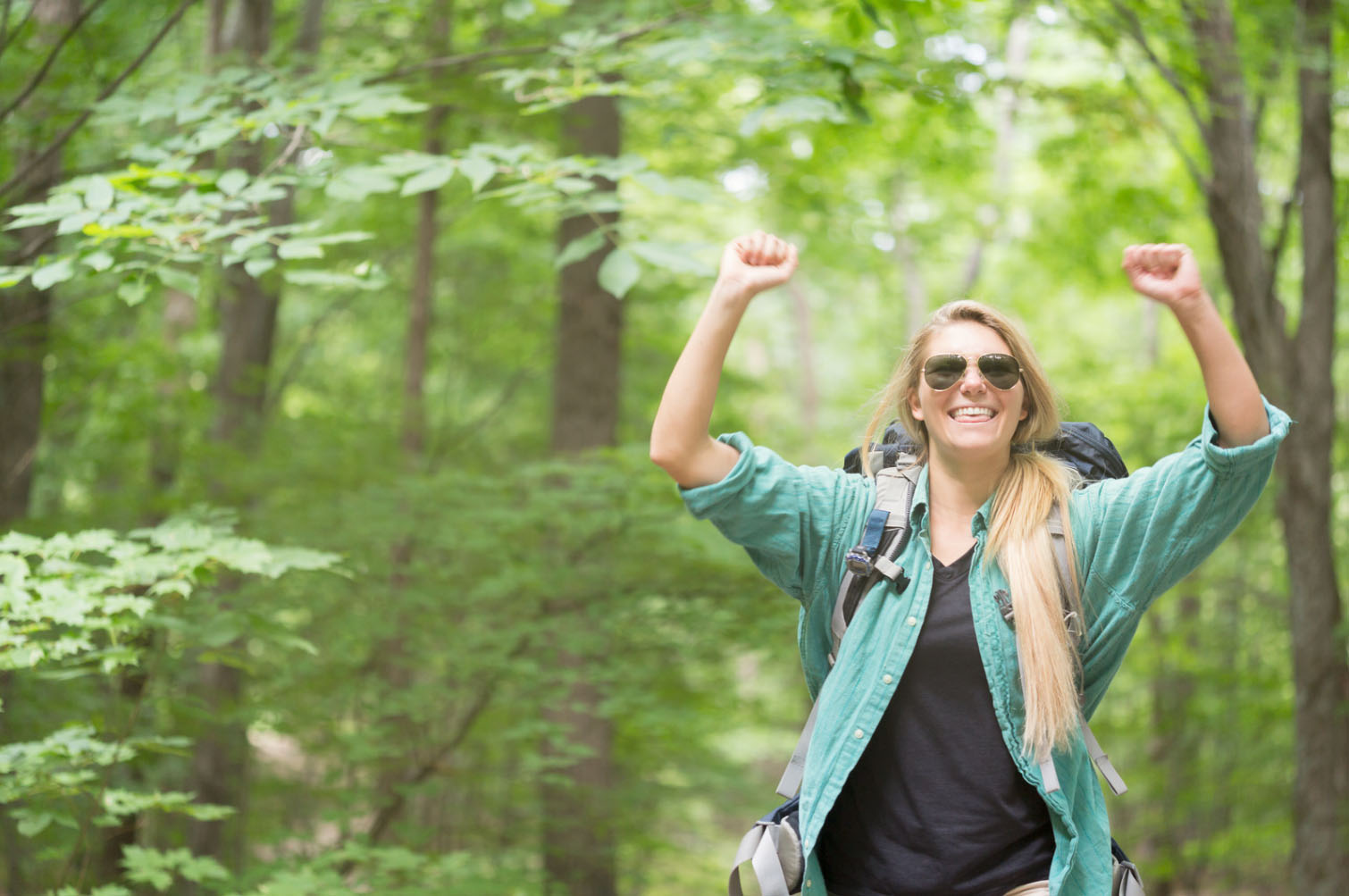 A woman with long hair and sunglasses celebrates joyfully on a hiking trail, raising her arms in triumph against a lush green forest backdrop, embodying outdoor adventure.