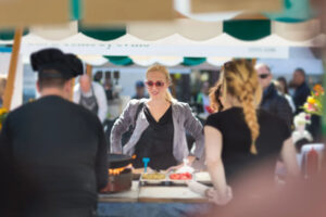 A woman with sunglasses stands at a food stall, engaging with customers while chefs prepare food at a lively outdoor market or food event.