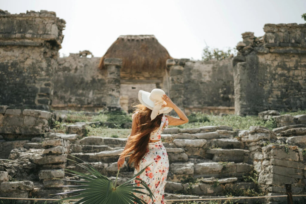 A woman wearing a white hat walks through the ancient ruins in Tulum, Mexico, with lush greenery and historical stone structures in the background.