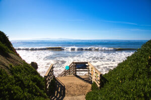 A wooden walkway leading to the beach with ocean waves crashing in the background under a clear blue sky.